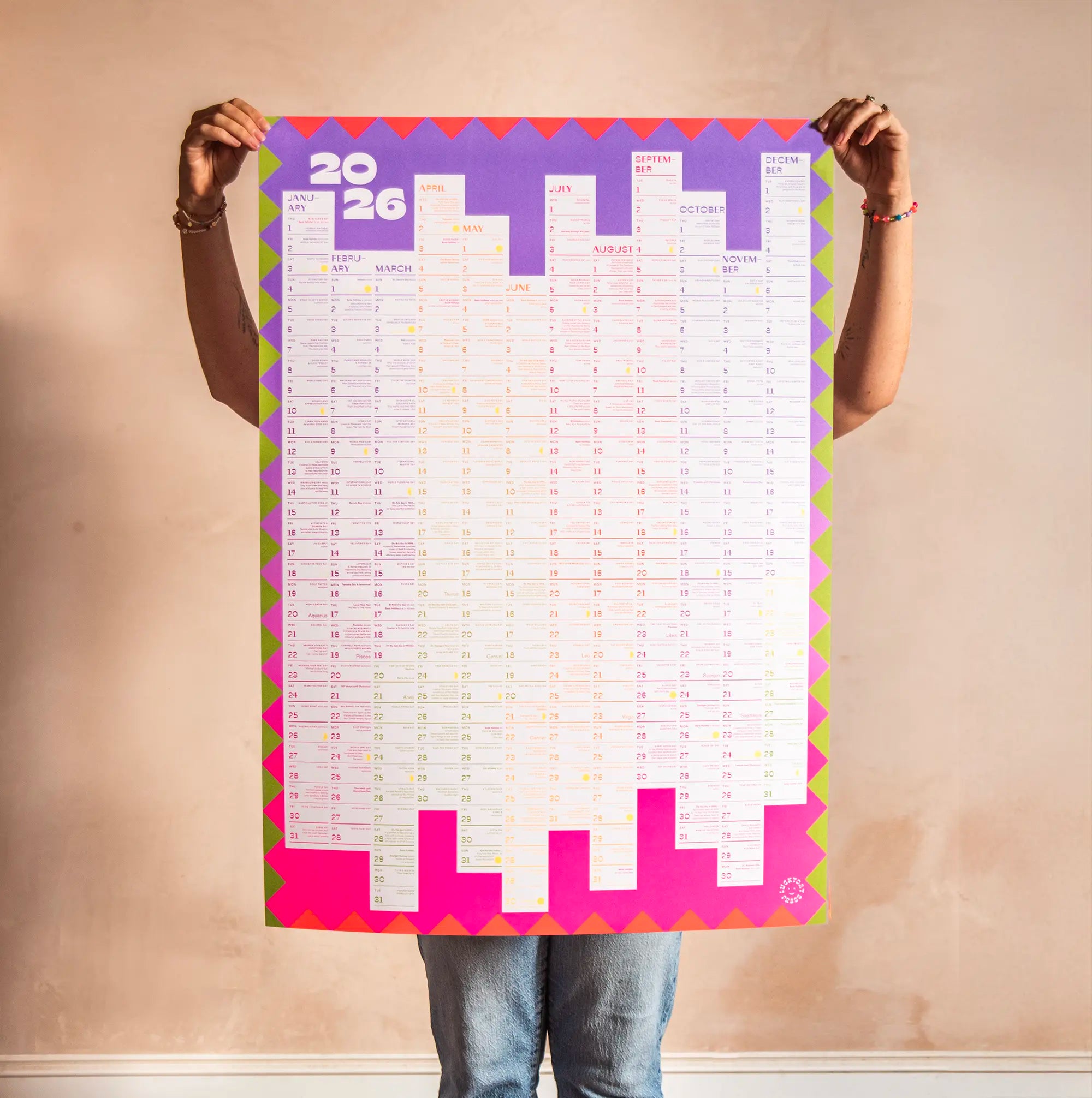 Person holding a large colorful kids A1 calendar poster for 2026 against a plain wall.