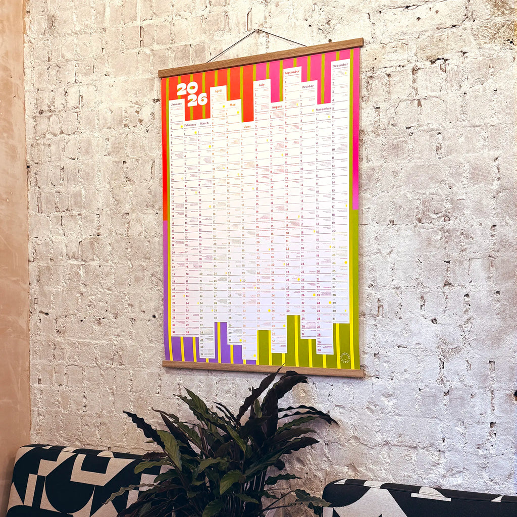 Colorful 2026 A1 calendar hanging on a textured wall with a plant in the foreground.