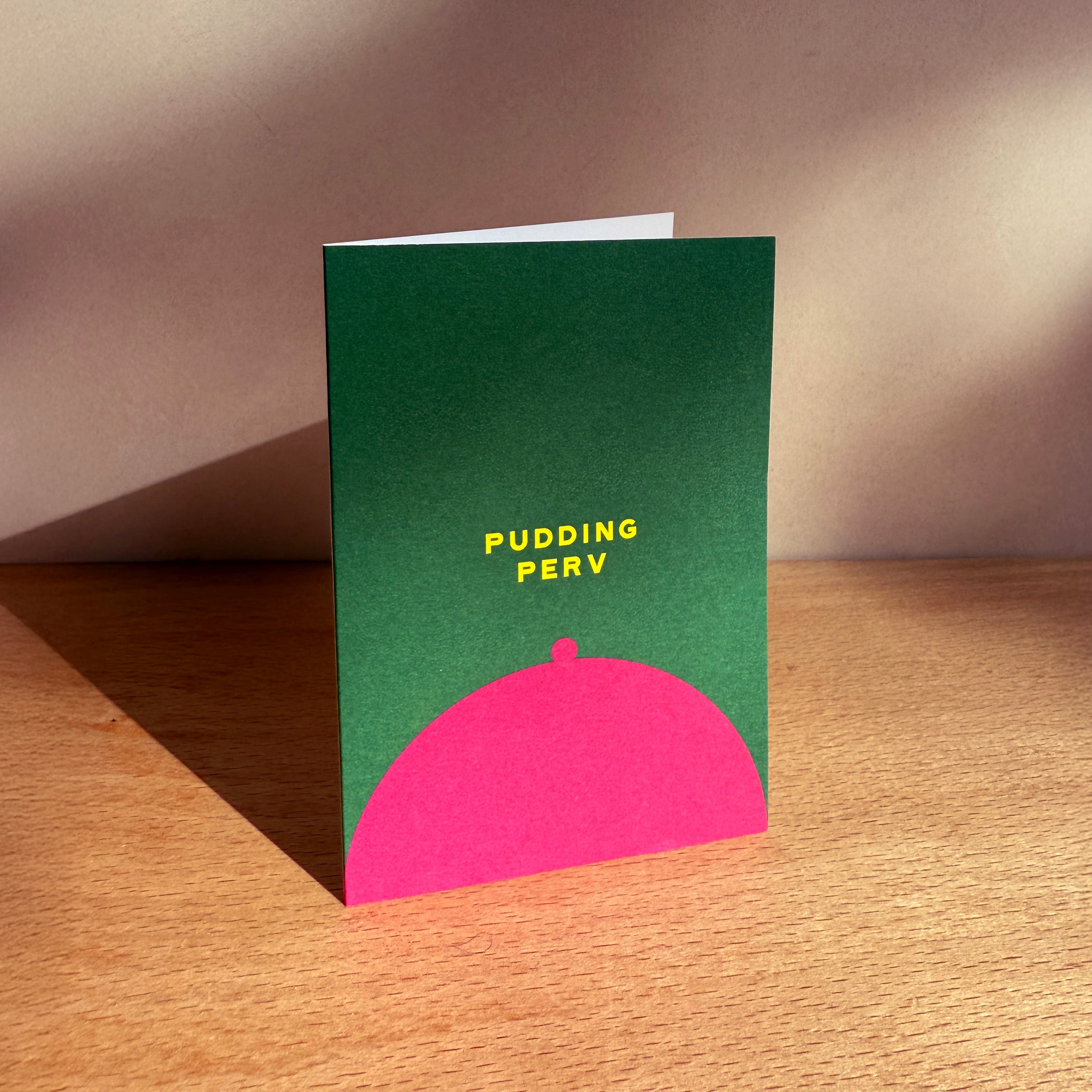 Green card with pink dome and yellow text 'PUDDING PERV' on a wooden surface.